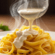 Um prato de macarrão fettuccine sendo coberto por um molho branco cremoso e fumegante, pronto para ser servido.